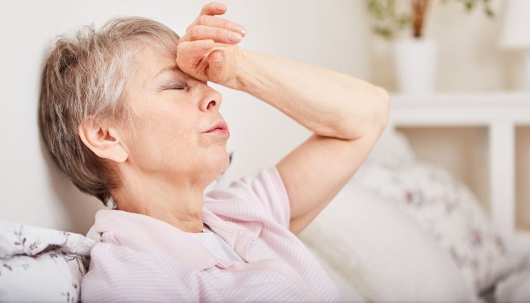 An older woman sitting on a couch with one hand on her forehead, as if she is nauseous.