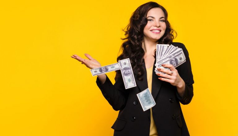 A woman in a black blazer holding a lot of $100 bills. She is standing in front of a bright yellow wall.